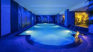 Piscina interior moderna com iluminação azul no teto e paredes, rodeada por piso ladrilhado e área de estar em curva.