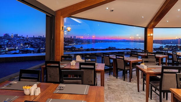 Un restaurante con mesas y sillas dispuestas para comer, amplias ventanas que ofrecen vista a un paisaje urbano y una masa de agua al anochecer.