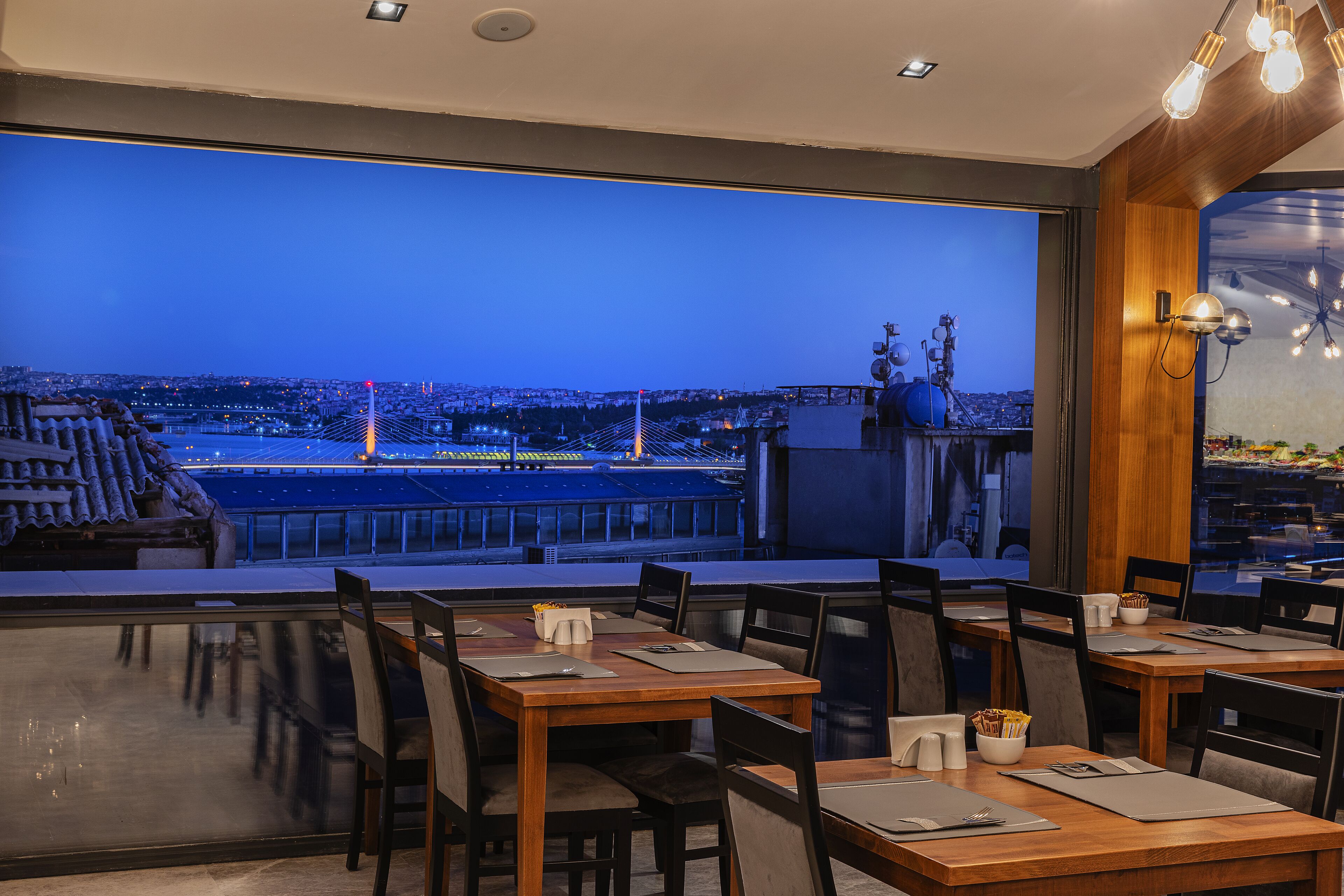 A restaurant with tables and chairs, a view of a cityscape and a bridge, and a wooden ceiling.
