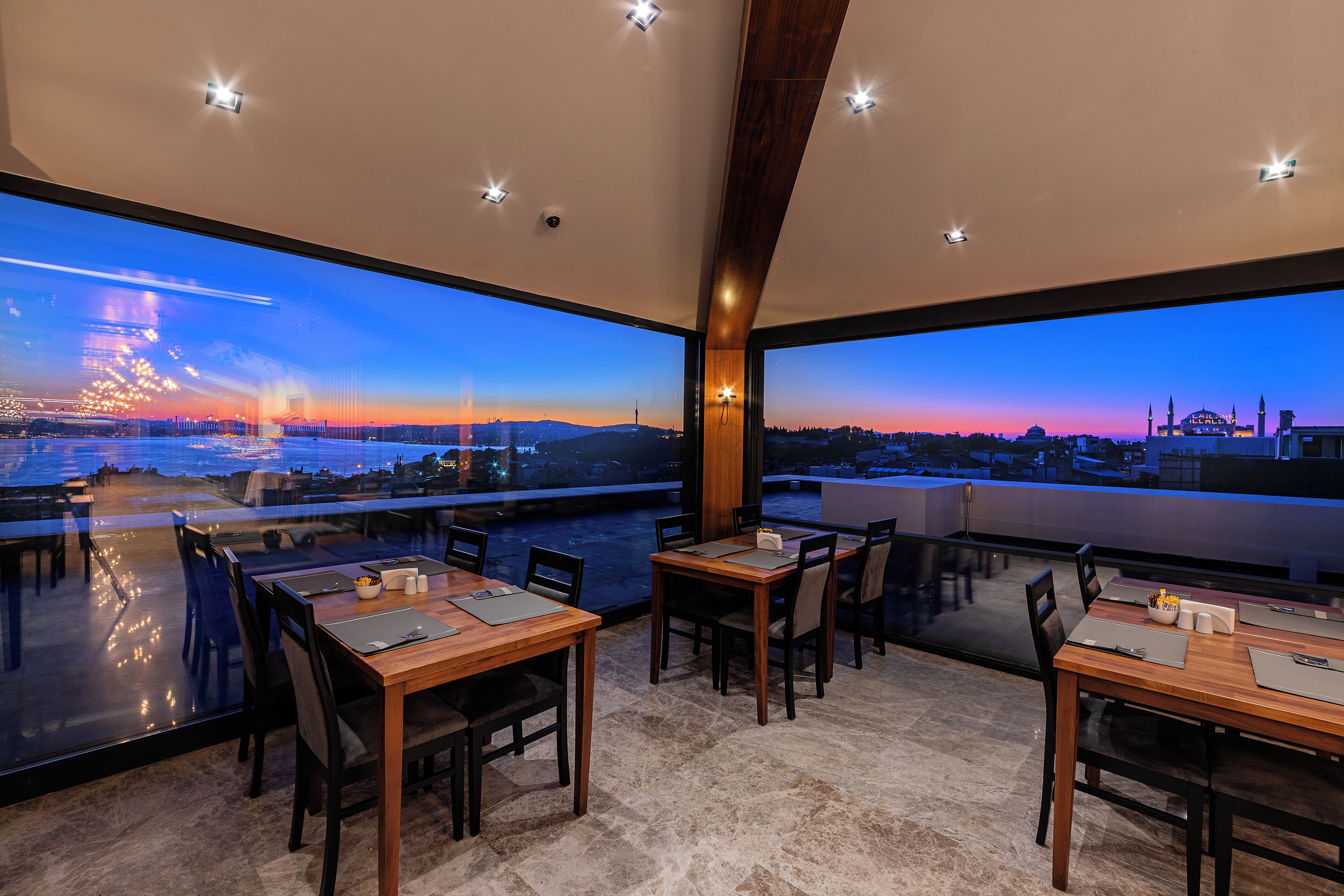 Ein Restaurant mit großen Fenstern, Holztischen und -stühlen mit Blick auf eine Stadtlandschaft bei Sonnenuntergang.