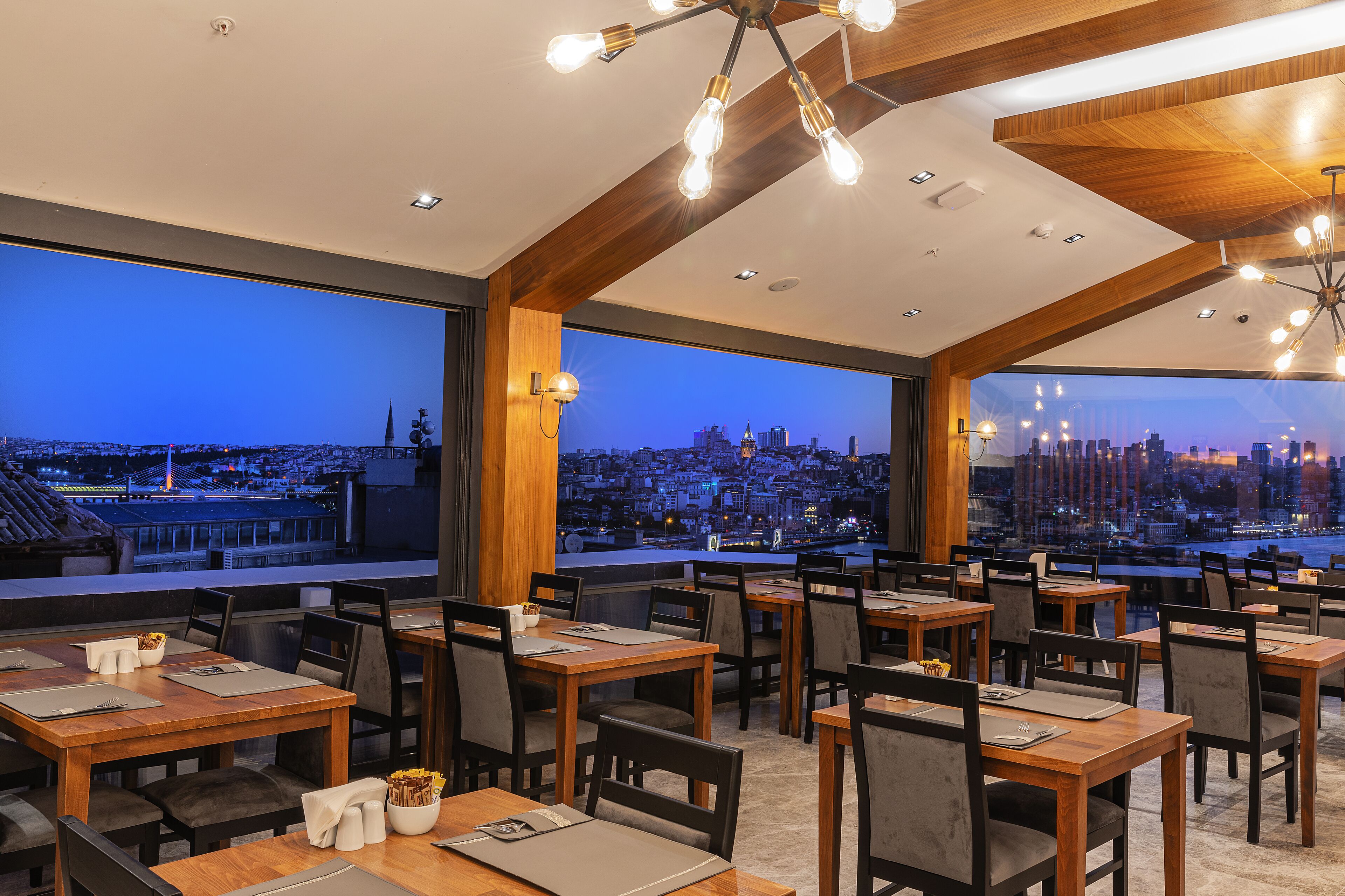 Ein Restaurant mit Tischen und Stühlen, großen Fenstern mit Blick auf die Stadt und einem modernen Deckenbau mit freiliegenden Balken und Beleuchtung.