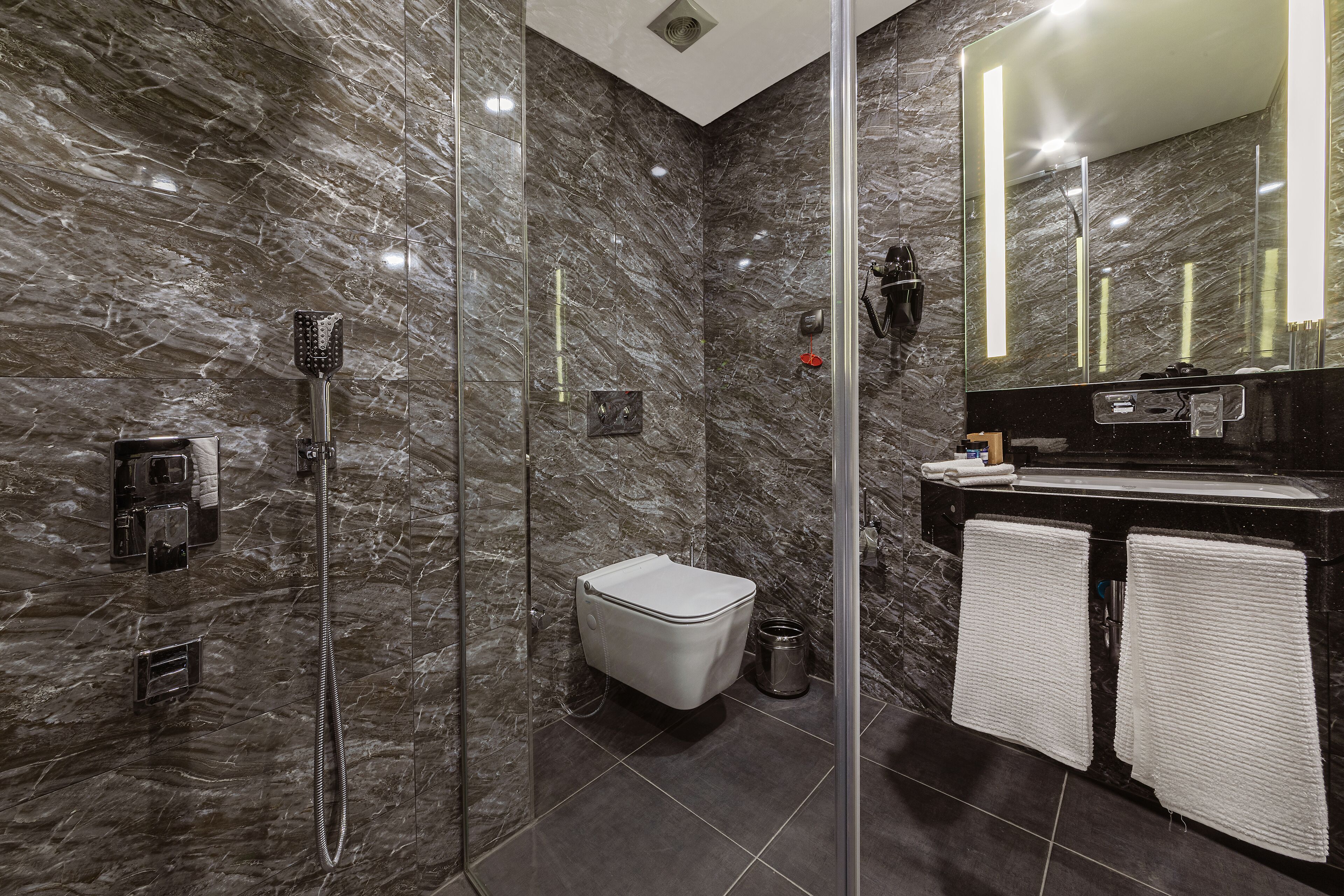 Un baño moderno con ducha de vidrio, inodoro blanco y lavamanos negro con dos toallas blancas.