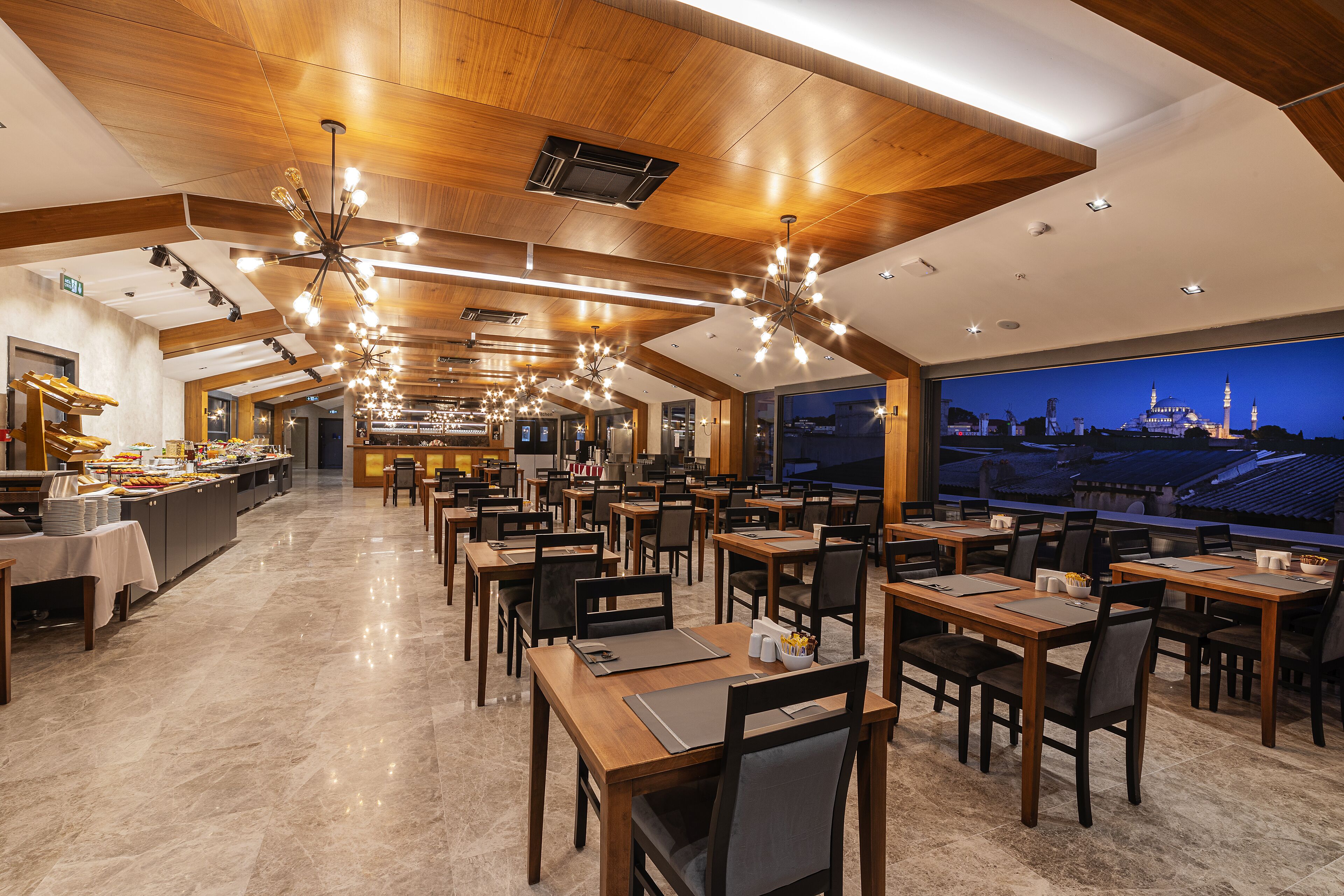 A spacious restaurant with high ceilings, modern chandeliers, and a view of a cityscape at dusk.