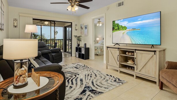 Ein Wohnzimmer mit einem großen Flachbildfernseher, einem Deckenventilator, einem schwarzen Sofa, einem Couchtisch mit einer Lampe und Blick auf den Strand durch die Glastür.
