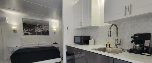 A compact hotel room with a bed, a small kitchenette, a microwave, a coffee maker, and a sink.