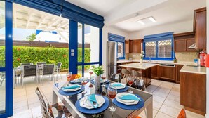 A modern kitchen with wooden cabinets, a built-in oven, and a dining area with a table set for four. - Villa Serena Springs by Ezoria Villas (Protaras)