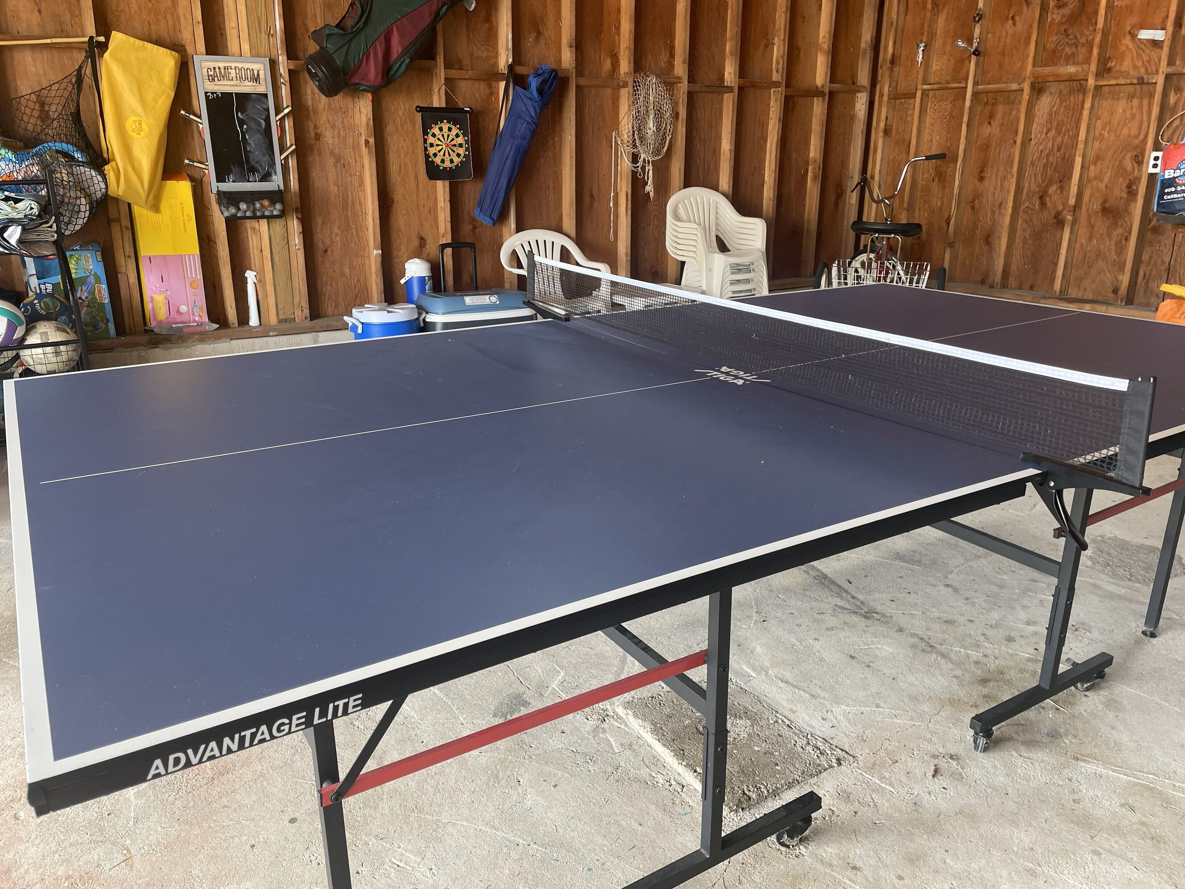 Une table de ping-pong avec un filet, entourée de divers équipements sportifs et d’un mur en bois.