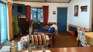 A cozy room with a wooden ceiling, a ceiling fan, a large window with curtains, a bed with a colorful blanket, a table with a tablecloth, and a sink with a soap dispenser. - Quaint Cottage on Swan Lake (Frankfort)
