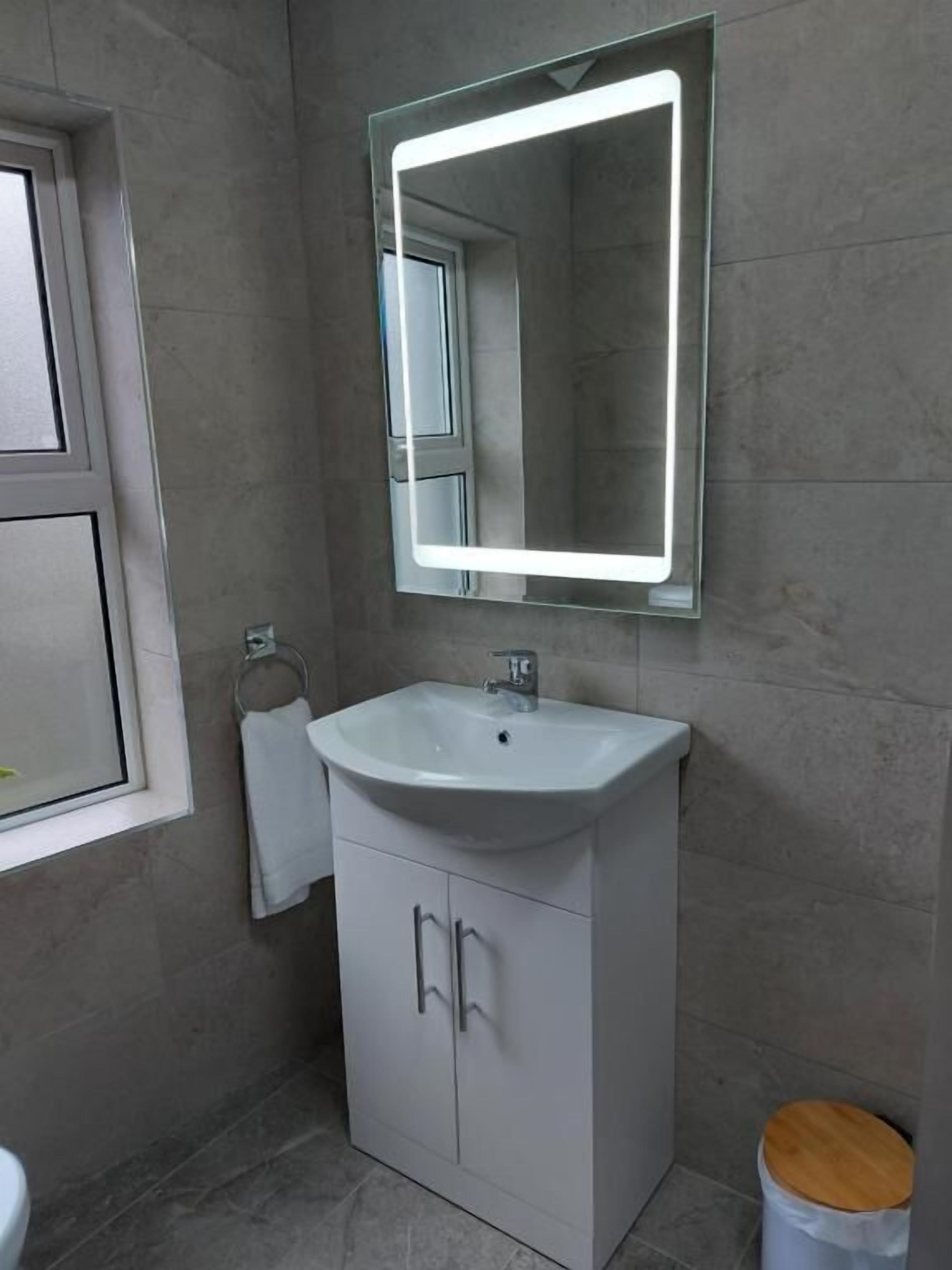 A bathroom with a white sink, a mirror with a light, and a wooden trash bin.