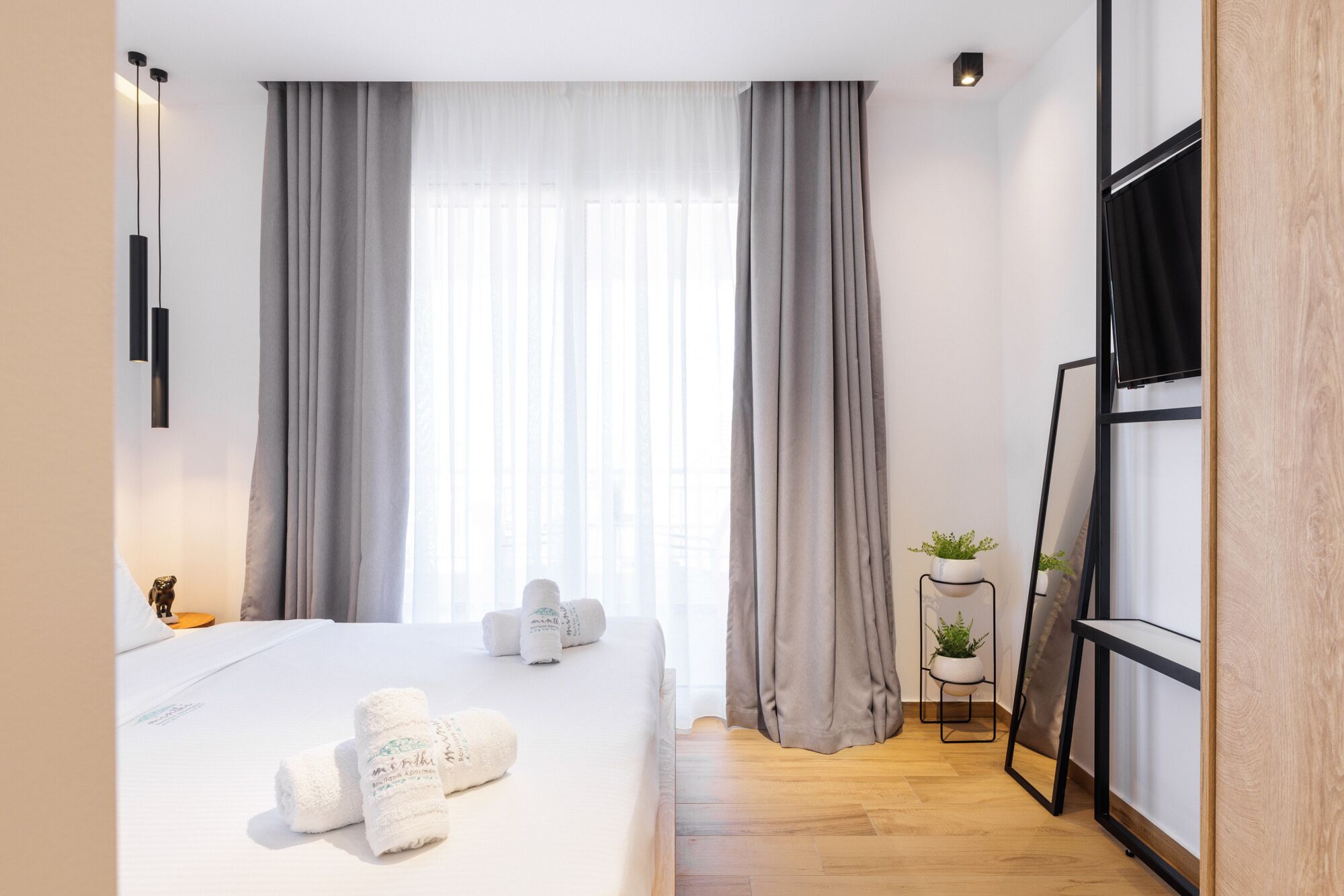 Una habitación de hotel moderna con una cama grande, cortinas grises, una mesita de noche con una planta y un espejo con marco negro.