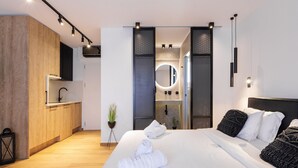 A modern hotel room with a wooden wardrobe, a built-in kitchen, a large bed with white linens, and a round mirror above the sink. - Kentro Luxury Suites (Kassandra)
