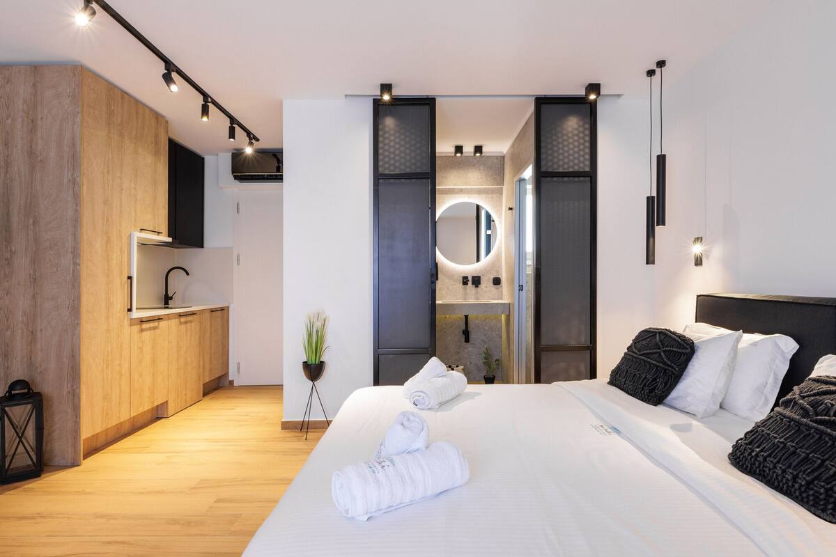 Ein modernes Hotelzimmer mit einem Holzschrank, einer integrierten Küche, einem großen Bett mit weißen Bettwäsche und einem runden Spiegel über dem Waschbecken.