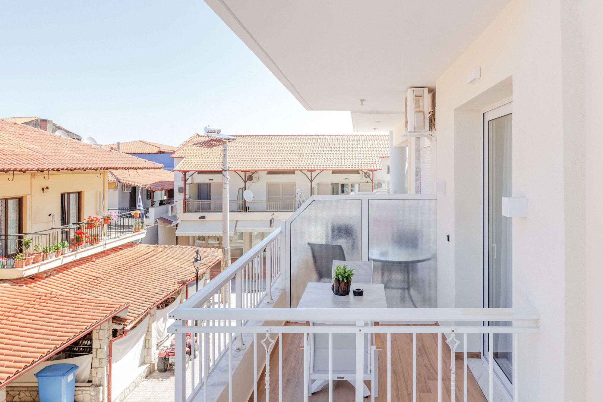 Ein Balkon mit weißer Brüstung, einem Tisch mit Topfpflanze und Blick auf benachbarte Gebäude mit roten Ziegeldächern.