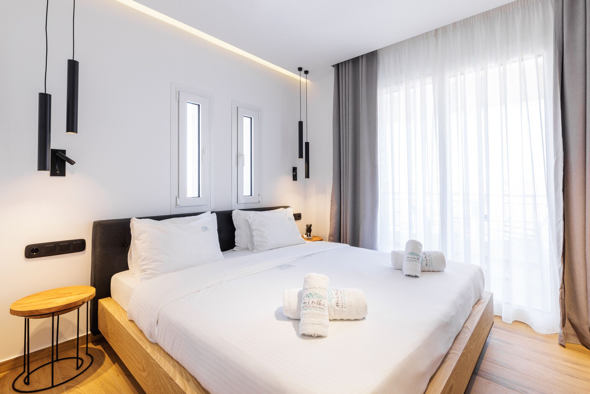 Une chambre d’hôtel avec un grand lit, deux tables de chevet, un petit tabouret en bois et une fenêtre avec des rideaux.