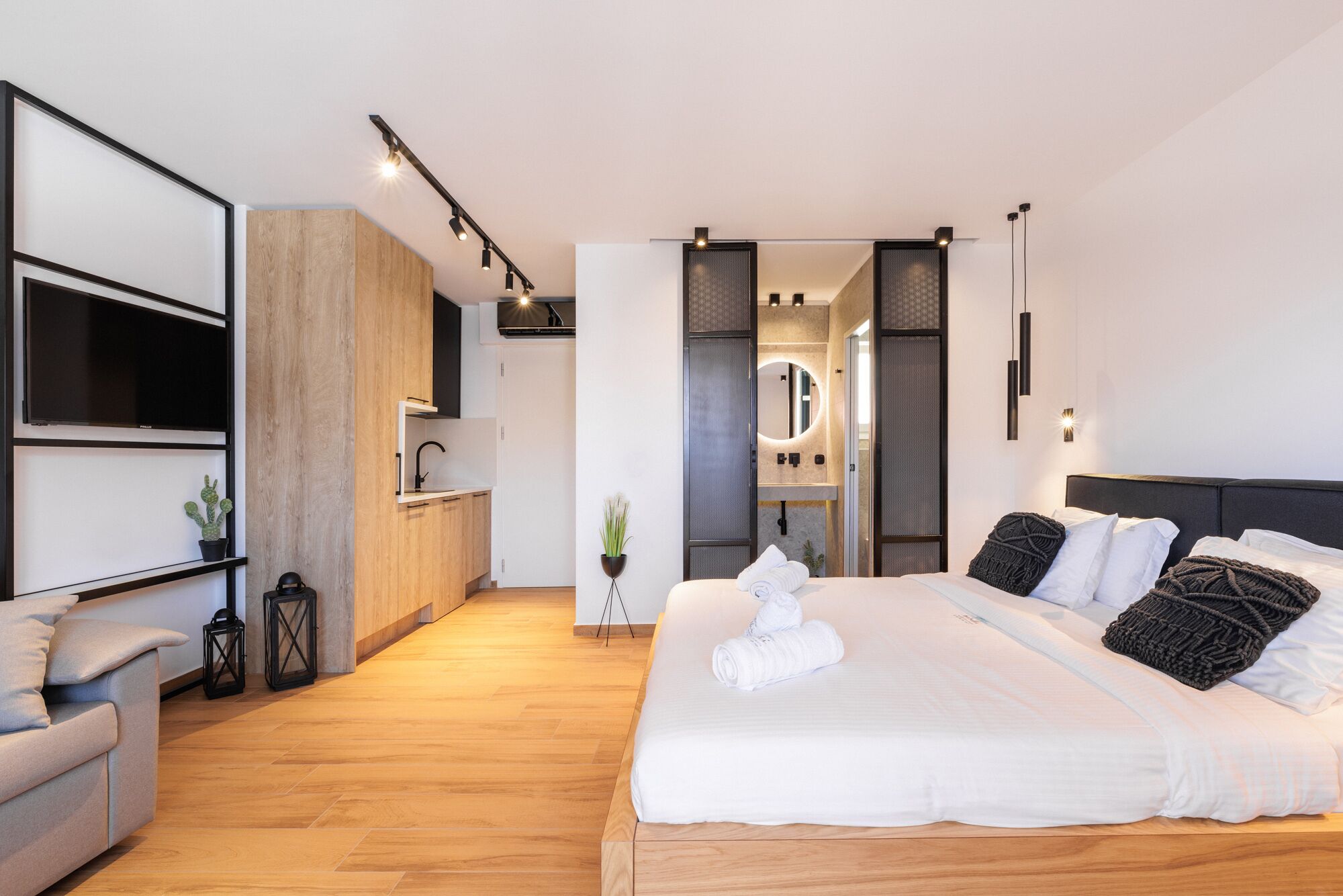 A modern bedroom with a large bed, a built-in wardrobe, a small kitchenette, and a bathroom with a mirror.