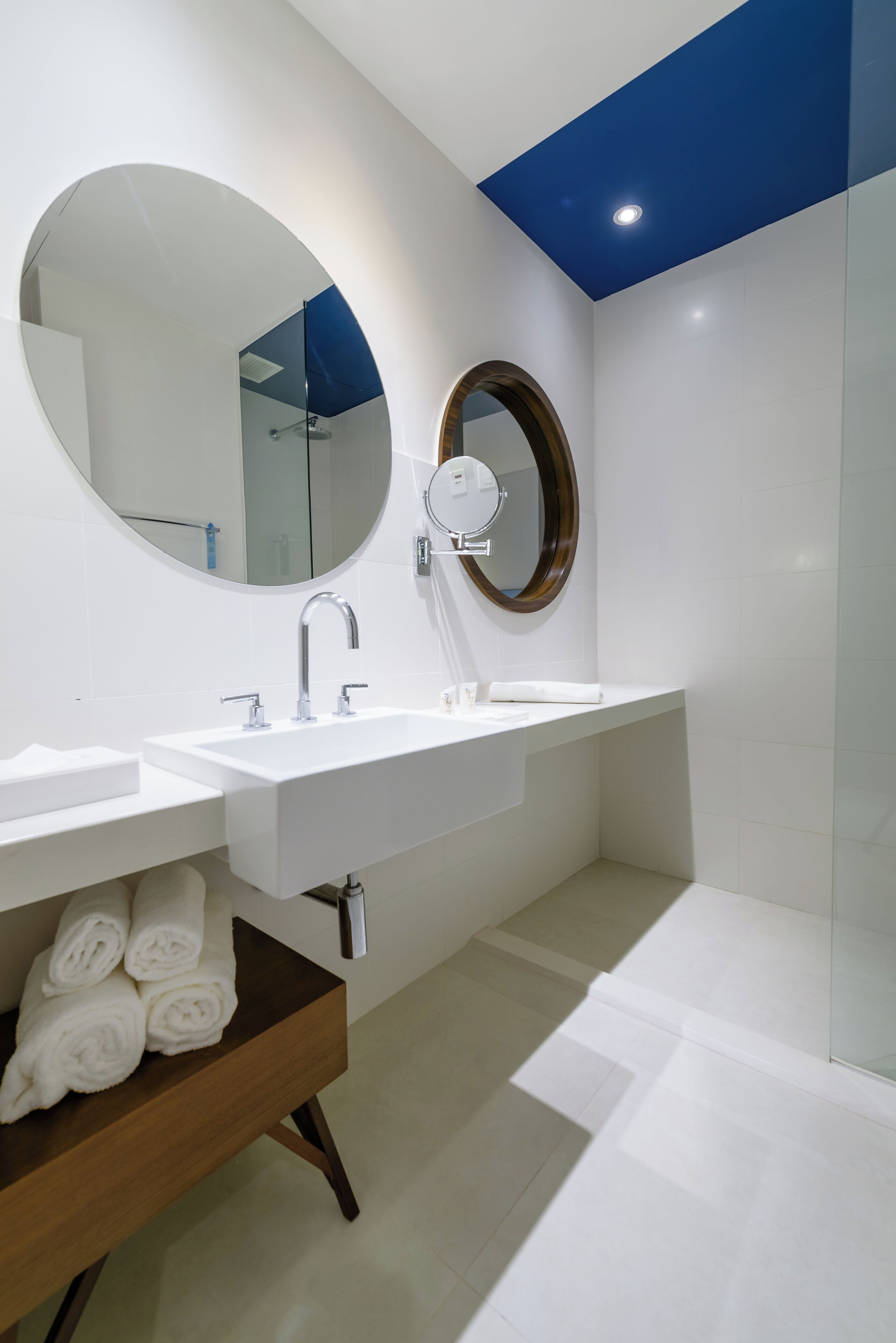 A modern bathroom with a white sink, a round mirror, and a wooden bench with towels.