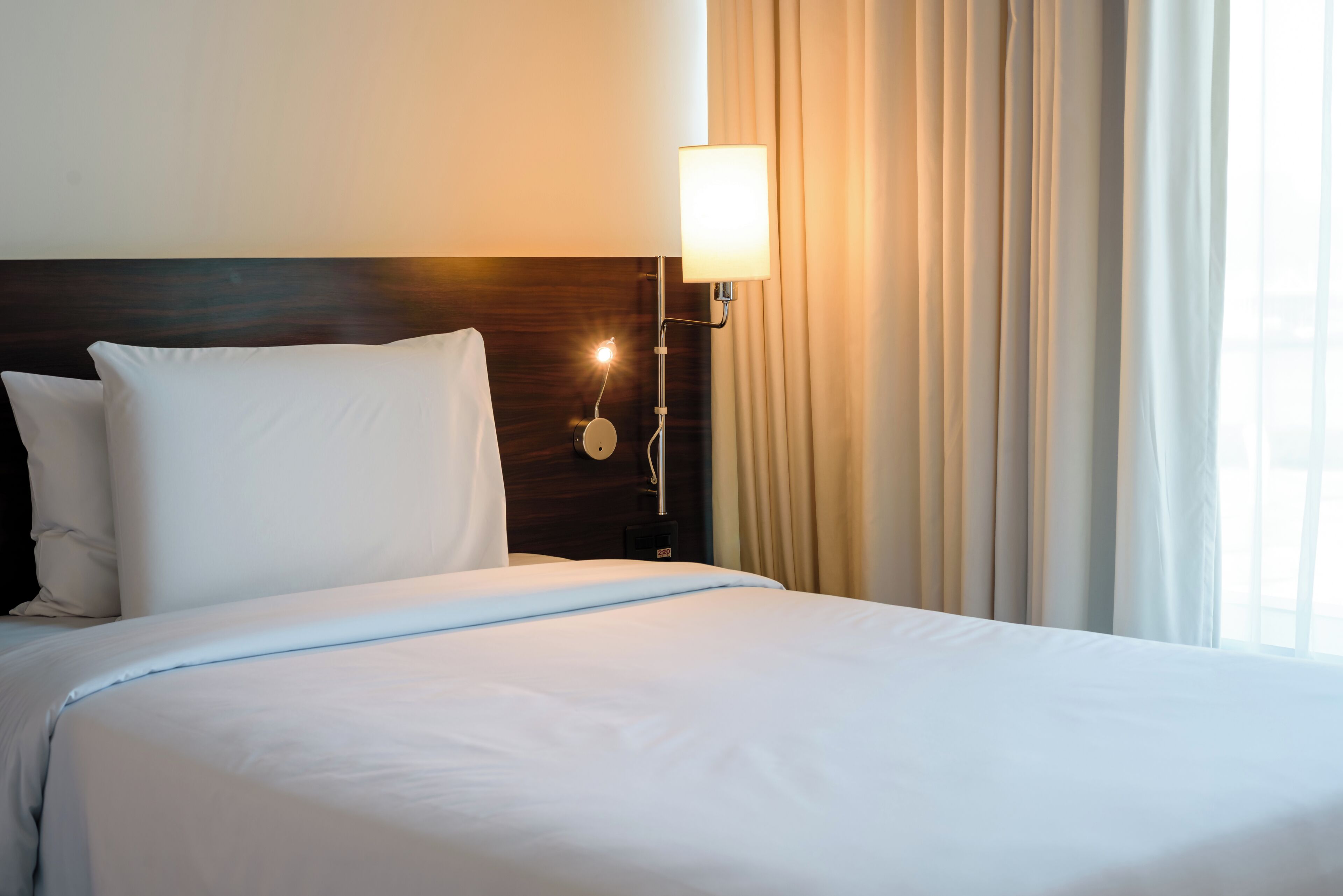Uma cama bem arrumada com lençóis brancos, cabeceira de madeira e luminária fixada na parede.