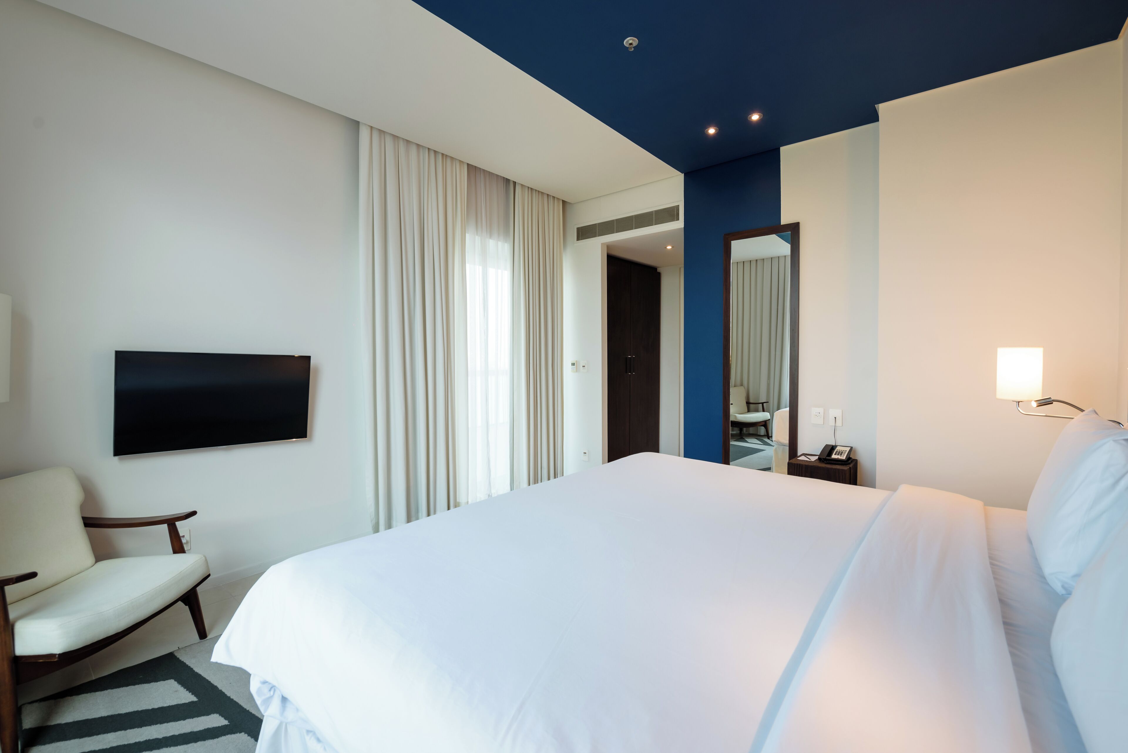 A hotel room with a large bed, a flat-screen TV mounted on the wall, a chair, and a desk with a lamp.