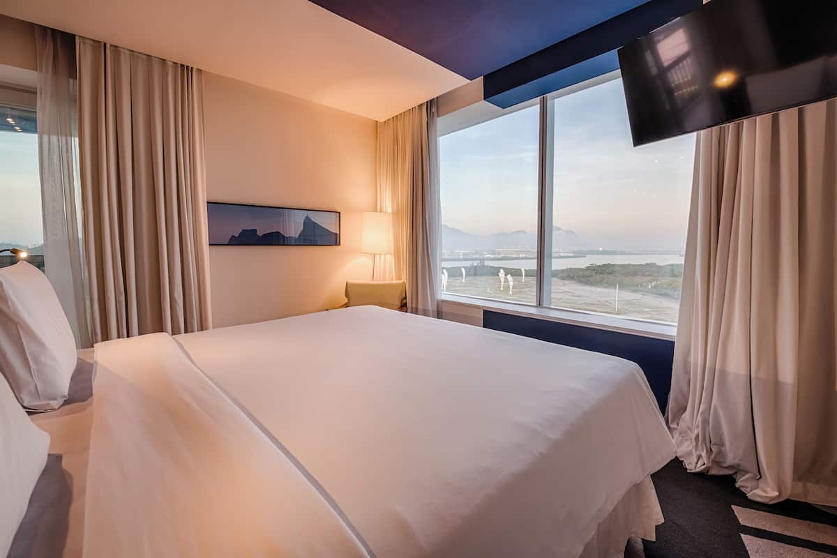 Een hotelkamer met een groot bed, een aan de muur bevestigde televisie, een raam met uitzicht op een brug en bergen, en een schilderij aan de wand.