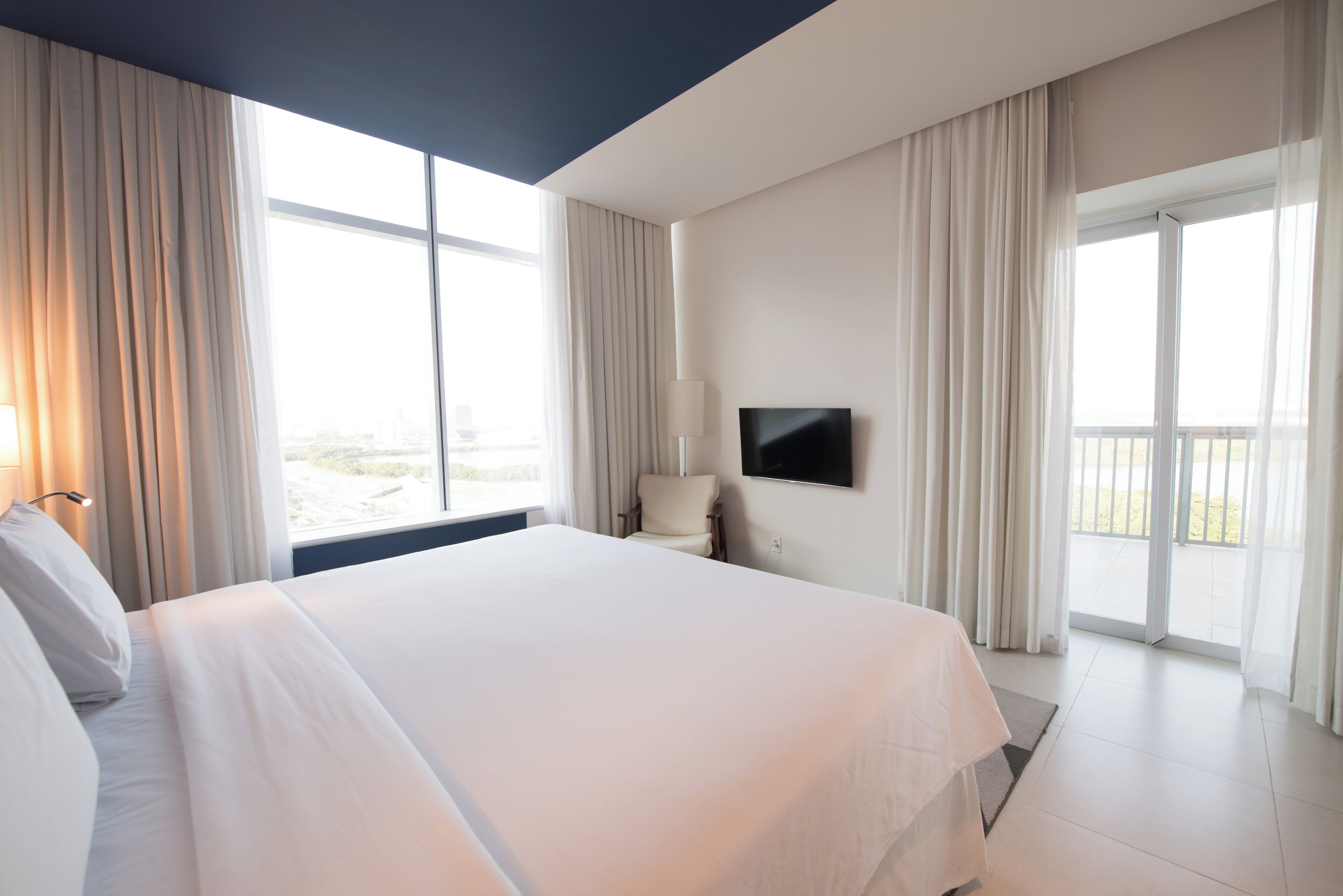 A hotel room with a large bed, a flat-screen TV mounted on the wall, and a balcony with curtains.
