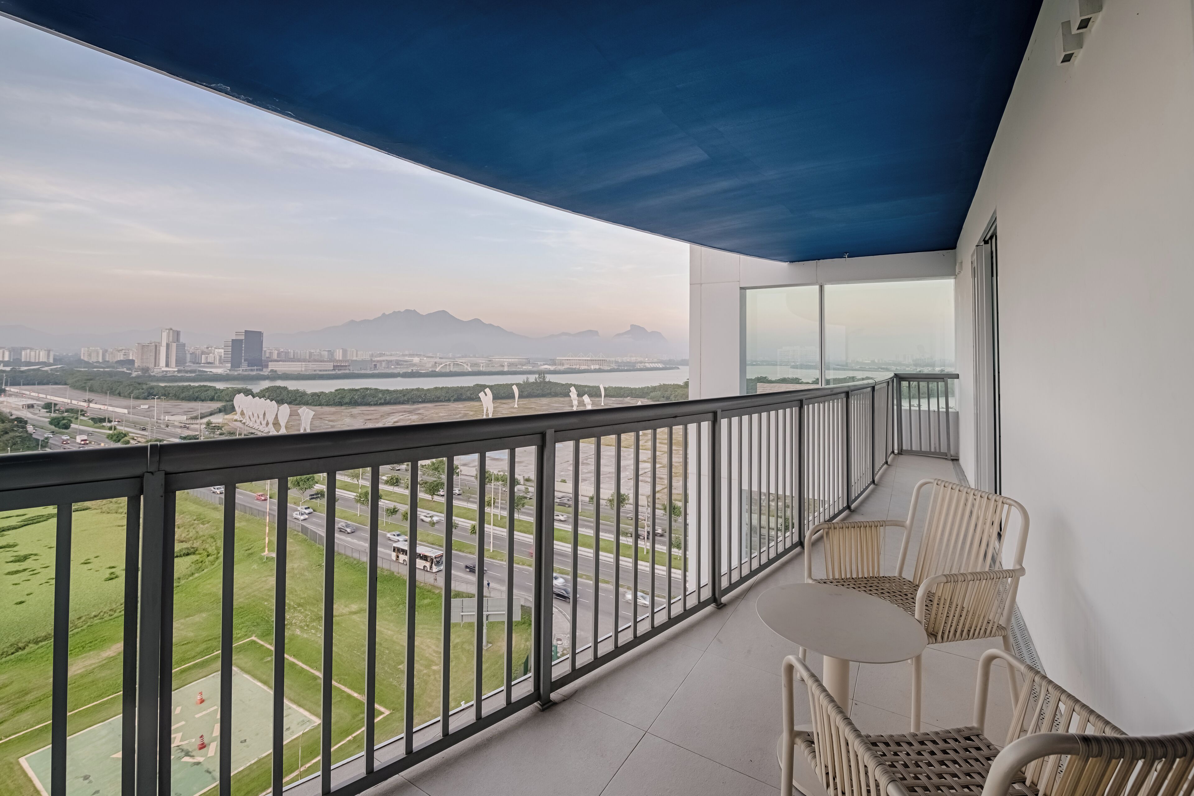 A balcony with a view of a cityscape, including buildings and a bridge. There are wicker chairs and a small table.