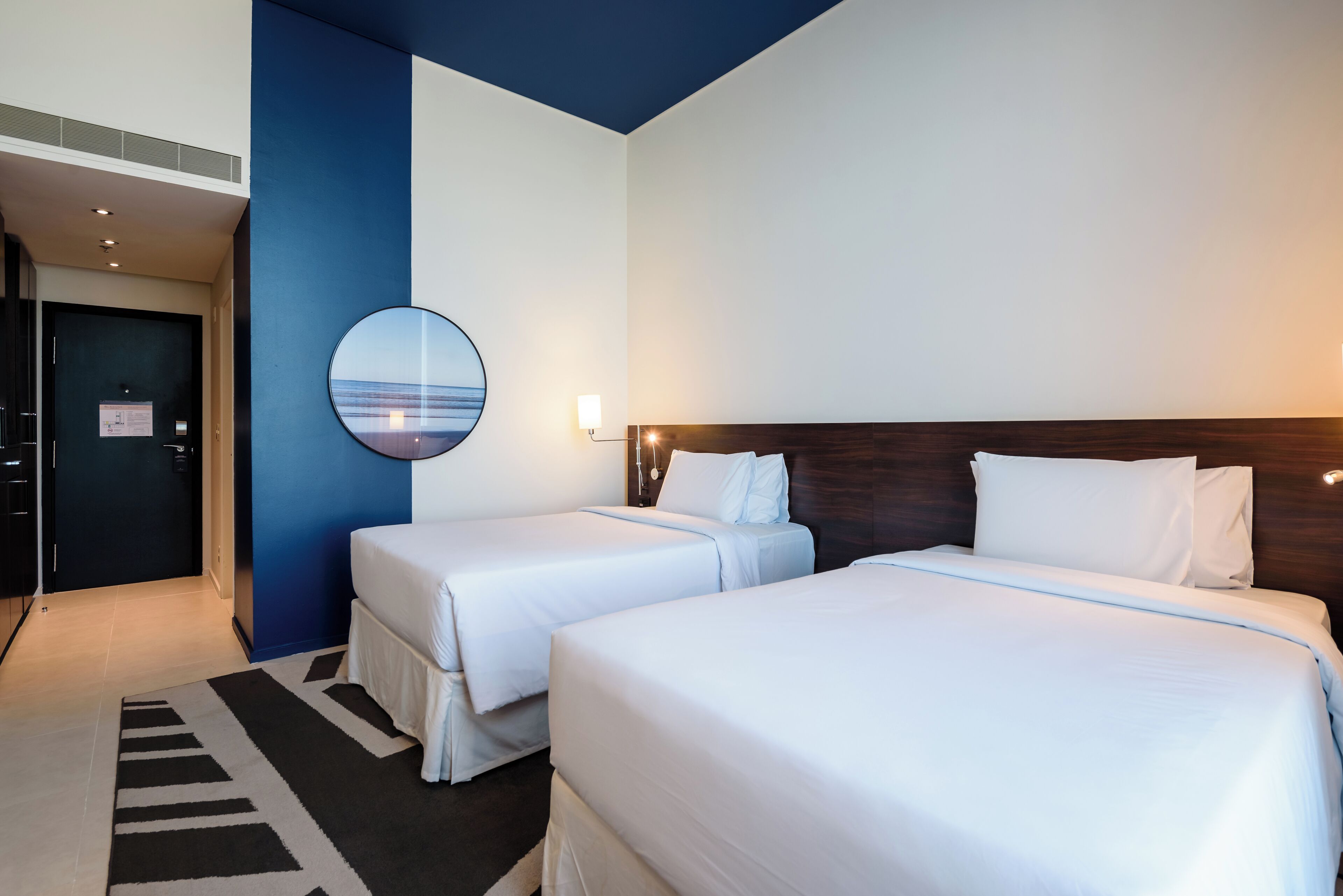 A hotel room with two single beds and a double bed, a round mirror, and a view of the sea through the window.