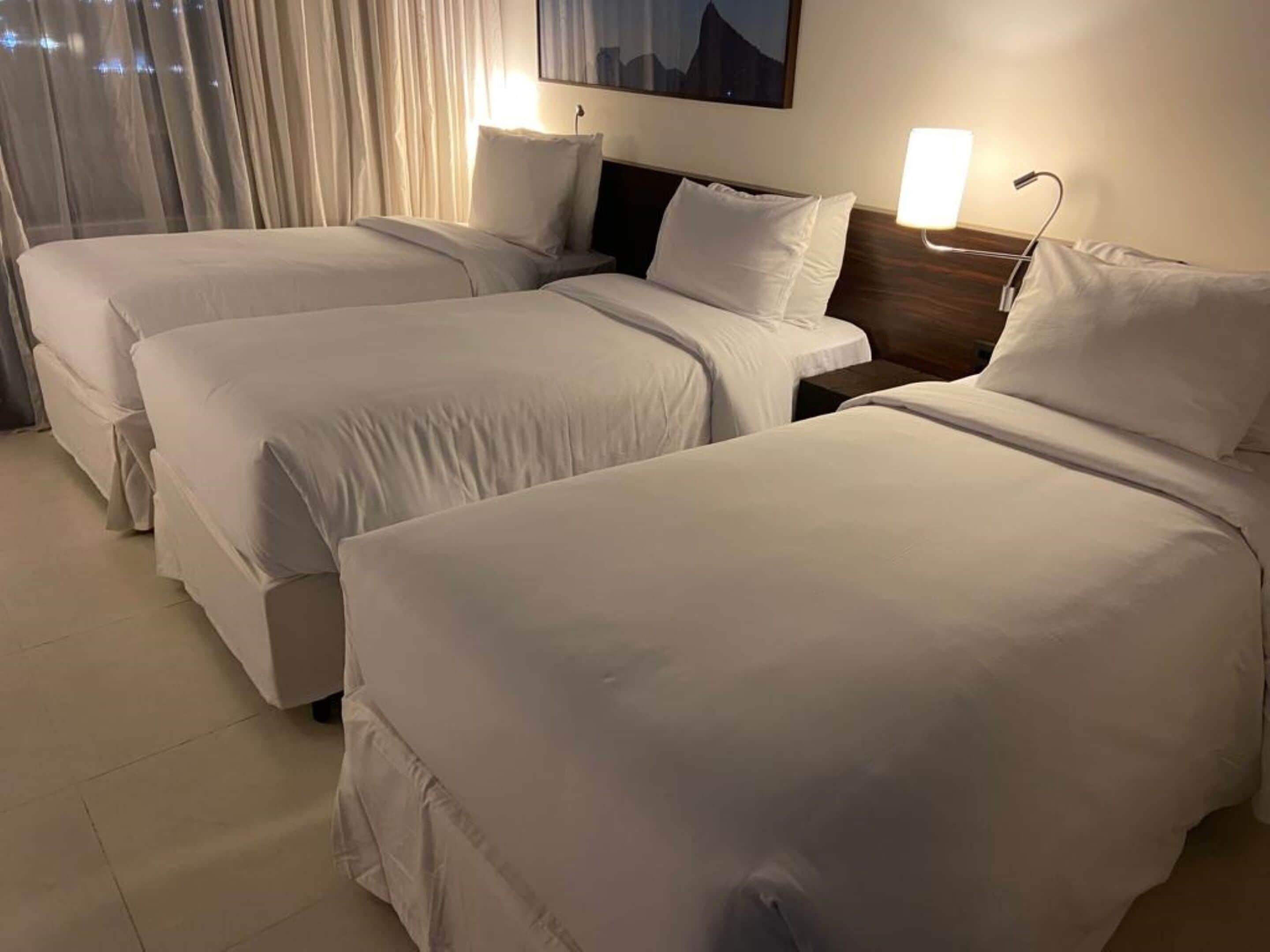 Une chambre d’hôtel avec trois lits simples, chacun doté de draps blancs et d’une tête de lit en bois. Il y a une table de chevet avec une lampe de chaque côté des lits.