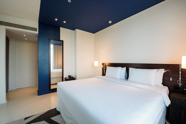 A modern hotel room with a large bed, a nightstand, a mirror, and a wall-mounted lamp.