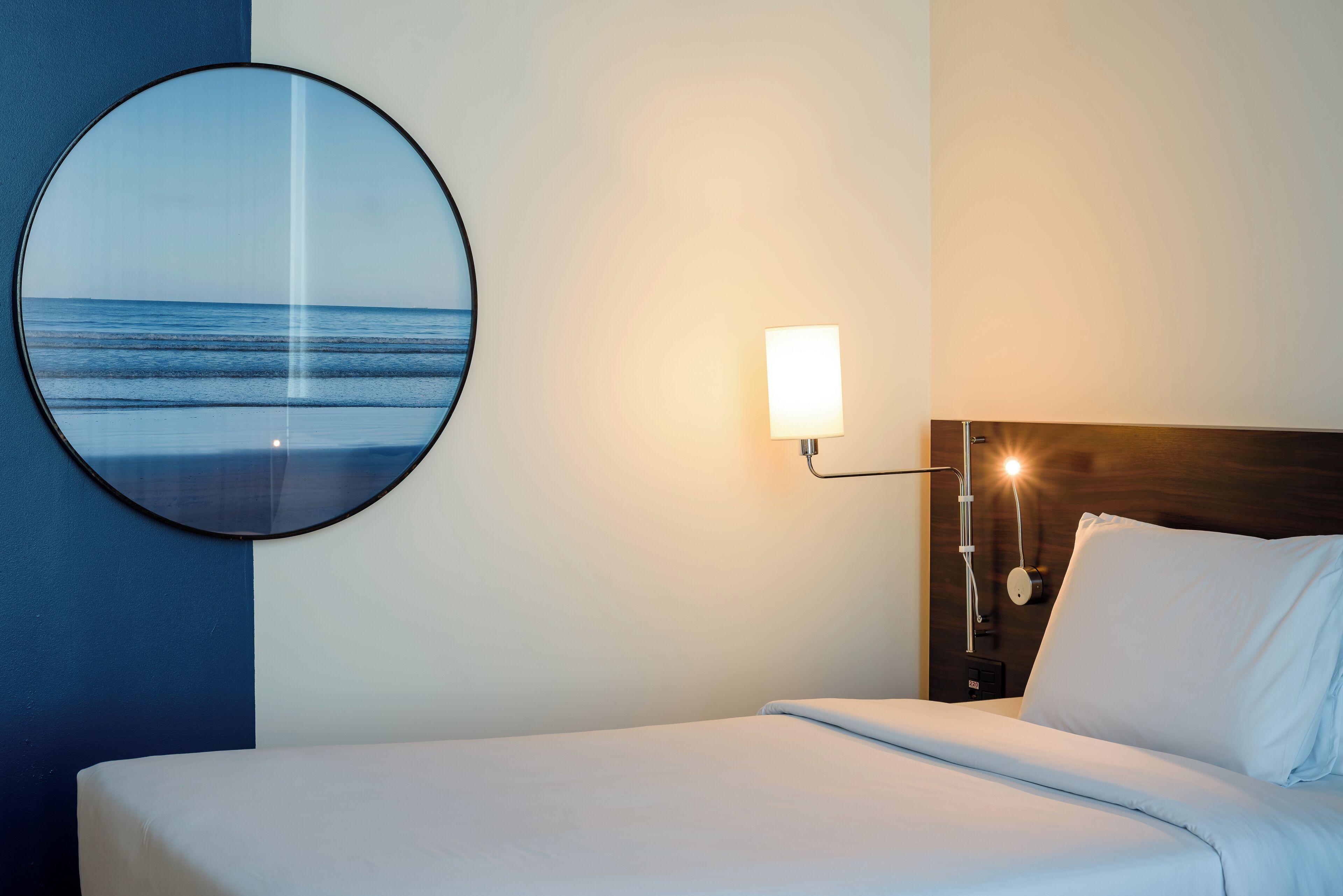 Habitació d'hotel amb un llit, un mirall rodó amb vista al mar, una taula de nit amb una làmpada i una meseta de nit amb un rellotge.