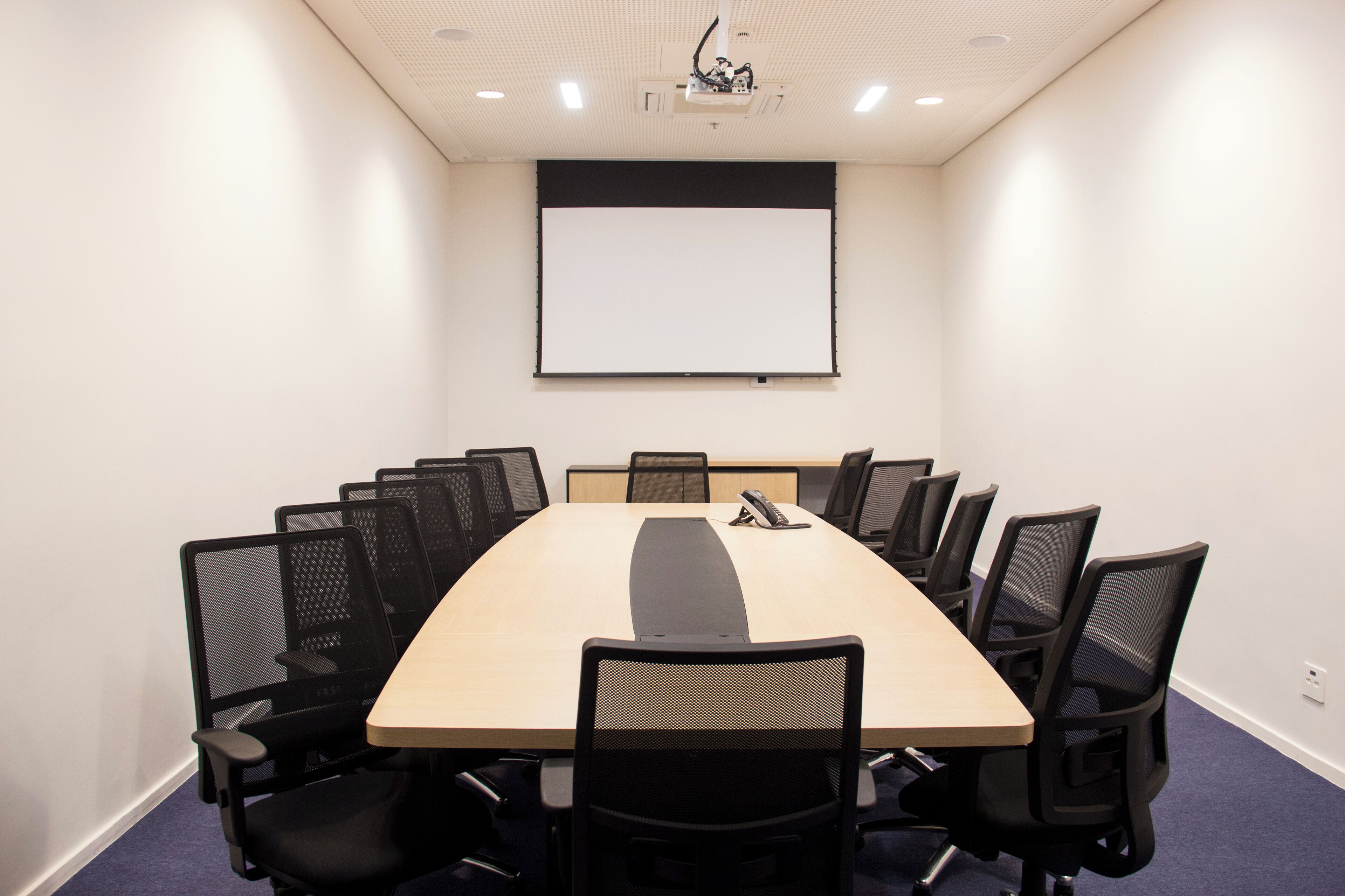 A conference room with a long table, black mesh chairs, a projector screen, and a whiteboard.