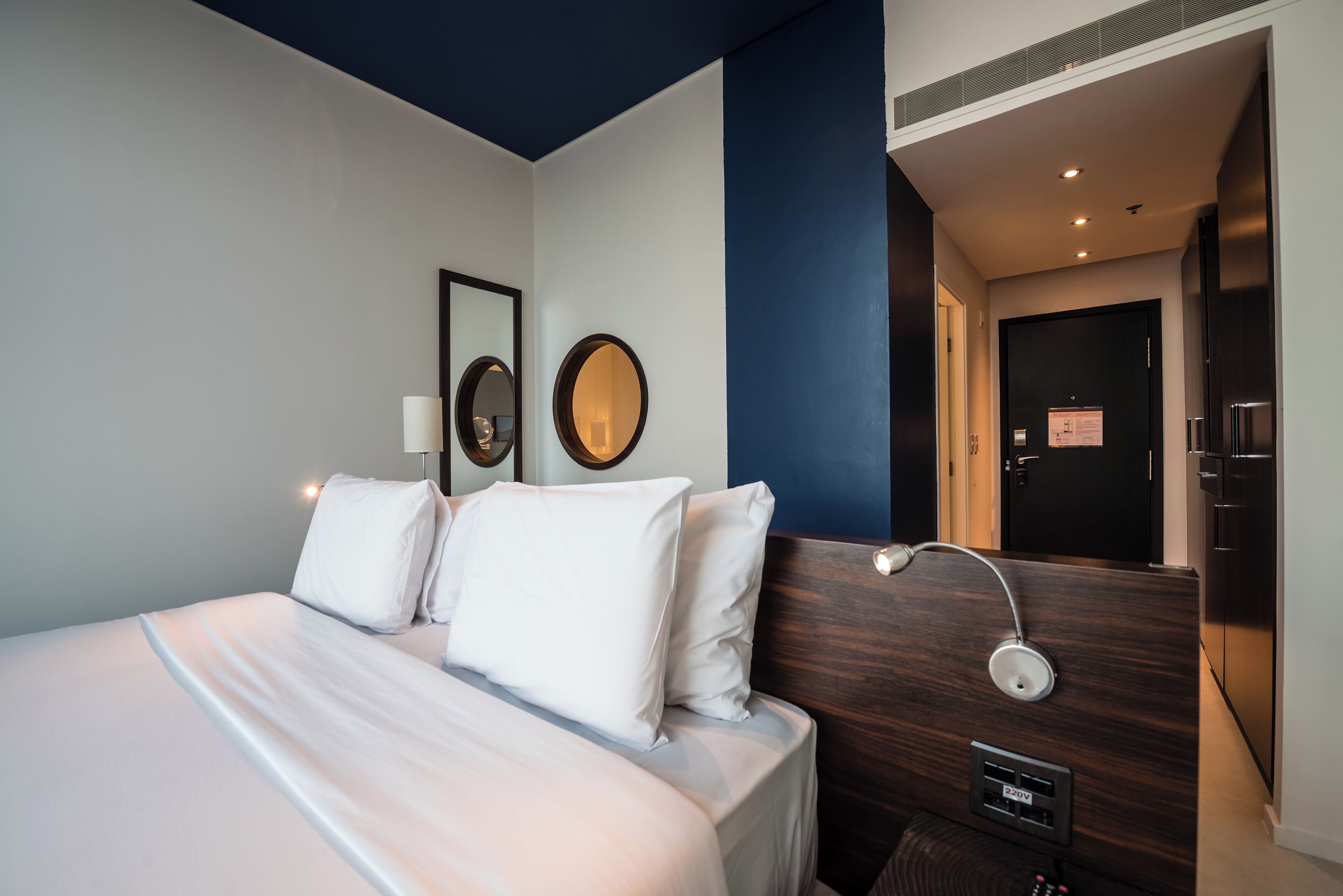 A hotel room with a bed, a mirror, a bedside lamp, and a door leading to a bathroom.