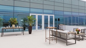 Un área de estar al aire libre con mesas y sillas, plantas en macetas y vistas a un edificio moderno con grandes ventanales.