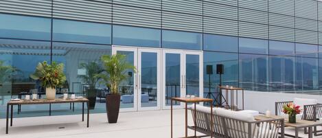 Un espace extérieur aménagé avec des tables et des chaises, des plantes en pot et une vue sur un bâtiment moderne doté de grandes fenêtres en verre.