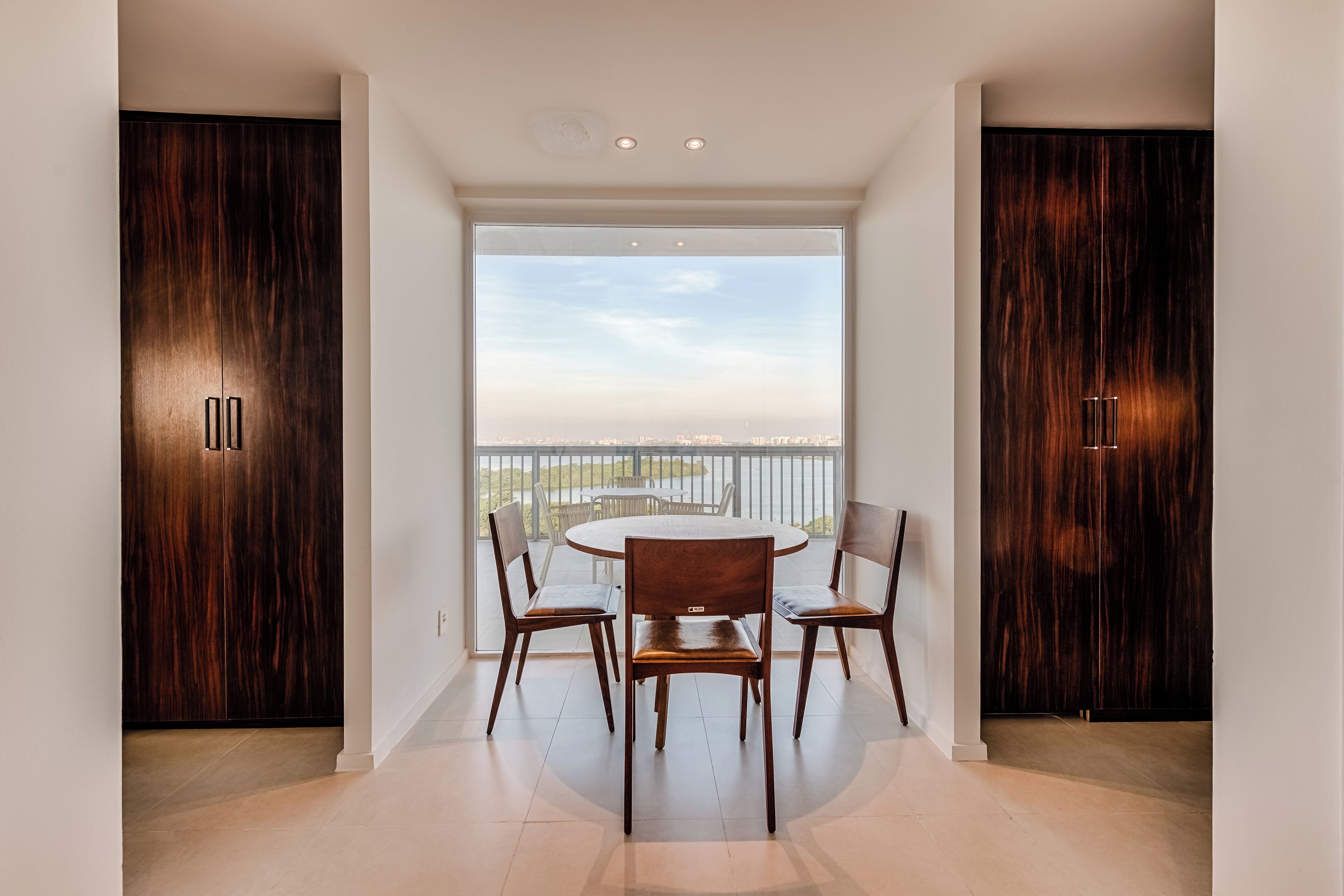 A modern dining area with a round table and four chairs, flanked by dark wooden cabinets, leading to a balcony with a view of the cityscape.