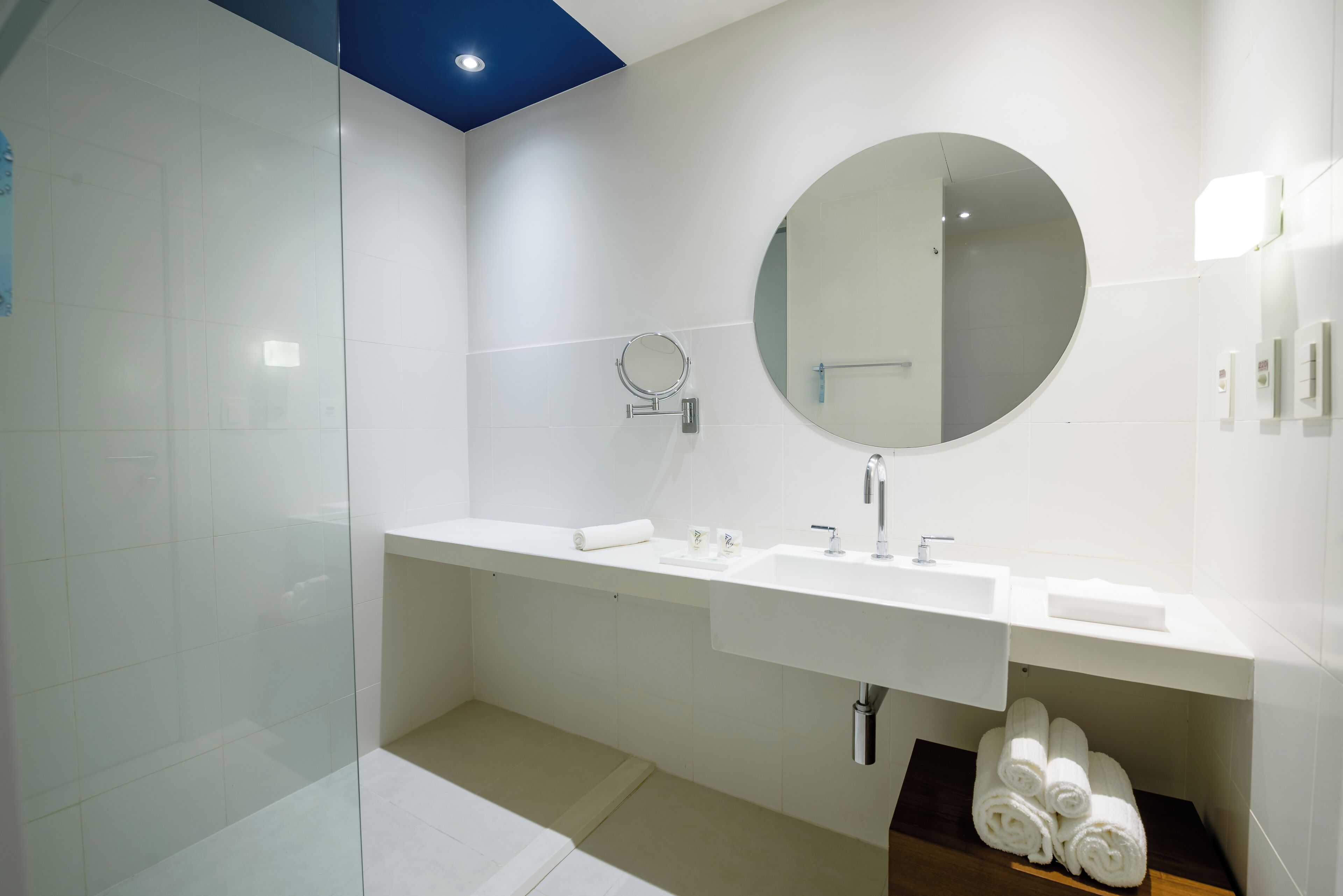 A modern bathroom with a glass shower enclosure, a white sink with a round mirror, and neatly rolled towels on a shelf.