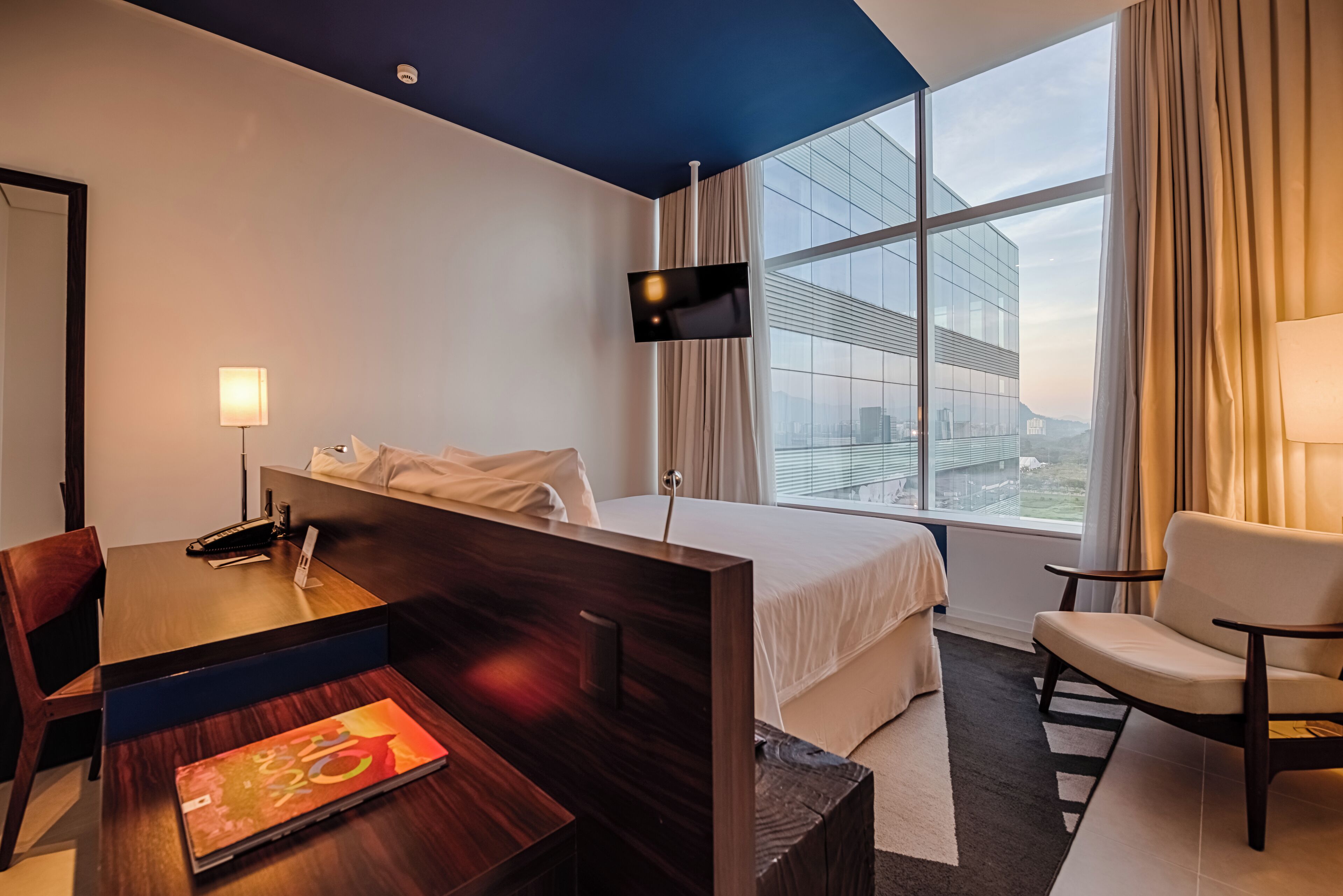 Une chambre d’hôtel équipée d’un lit, d’un bureau, d’une chaise, d’une télévision et d’une grande fenêtre donnant sur la ville.