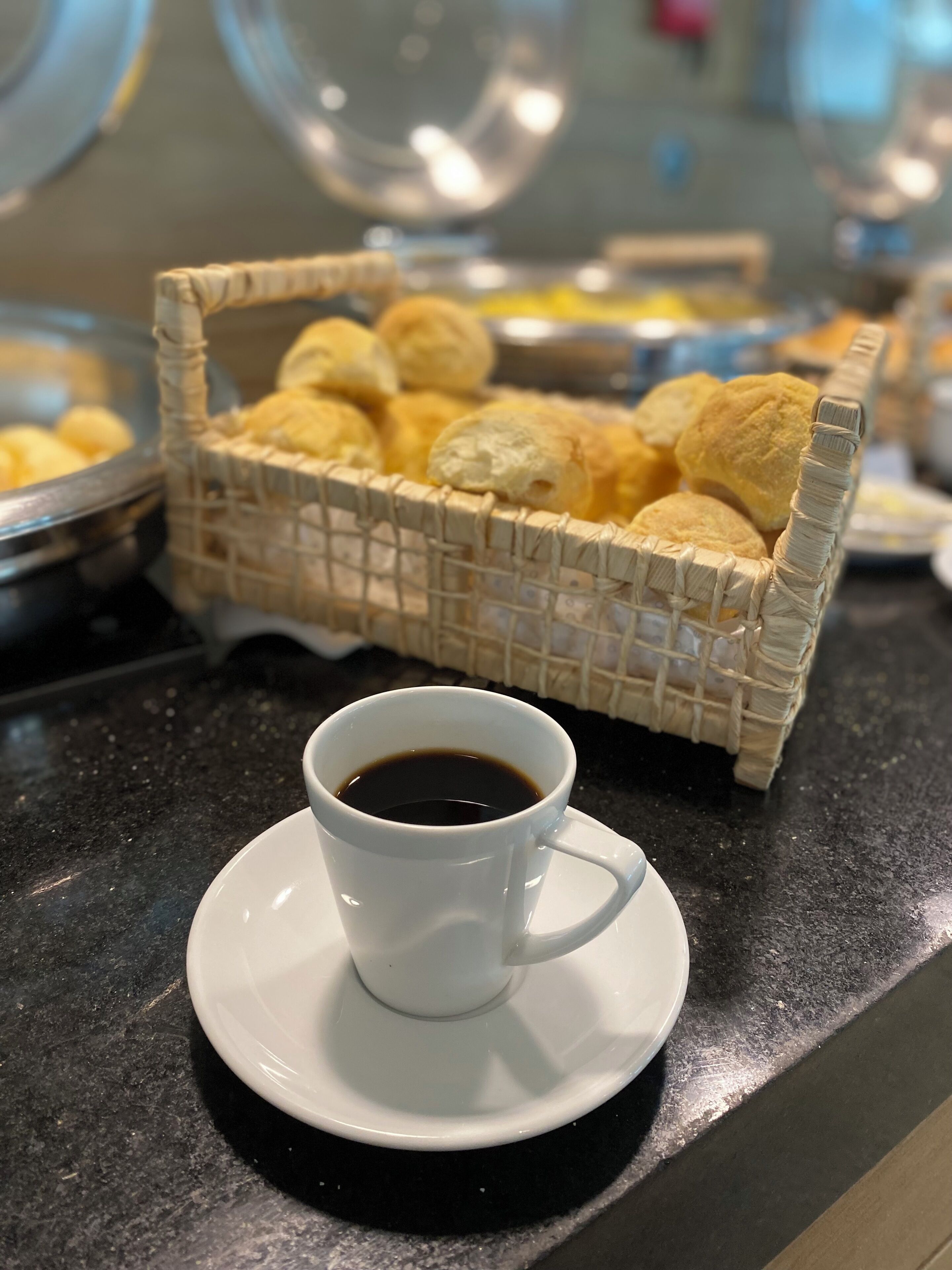 A white cup of coffee on a saucer, a wicker basket with pastries, and a buffet area in the background.