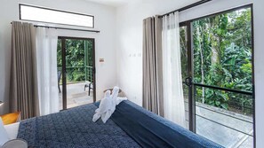 Quarto de hotel com cama, uma cadeira, uma mesinha e varanda com cortinas.