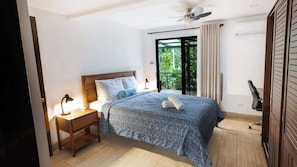 A bedroom with a bed, nightstand, desk, chair, and a view of greenery through a sliding door.