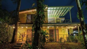 A modern two-story house with a balcony, surrounded by tropical vegetation.