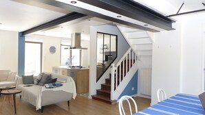 A modern living room with a sofa, a dining table, and a staircase leading to an upper level. - Cosy 3-star holiday home, 6 people (Ploeren)
