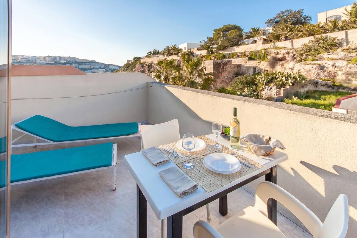 Una terraza con una mesa lista para comer, una silla de descanso y vistas a una colina con edificios y vegetación.
