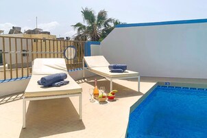Un área de piscina en la azotea con sillones de descanso, una mesa con bebidas y una piscina azul cristalina.