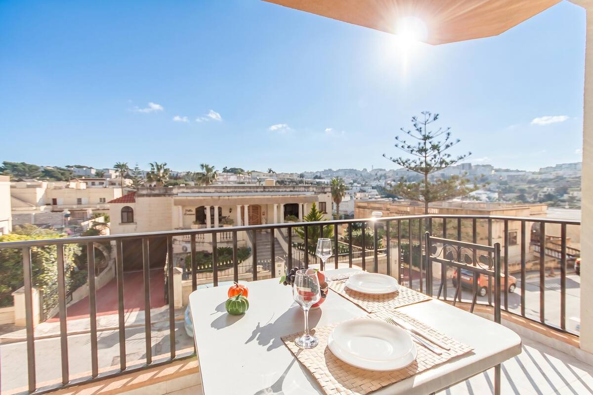 Un balcón con una mesa para dos, vistas despejadas de la ciudad y un día soleado.