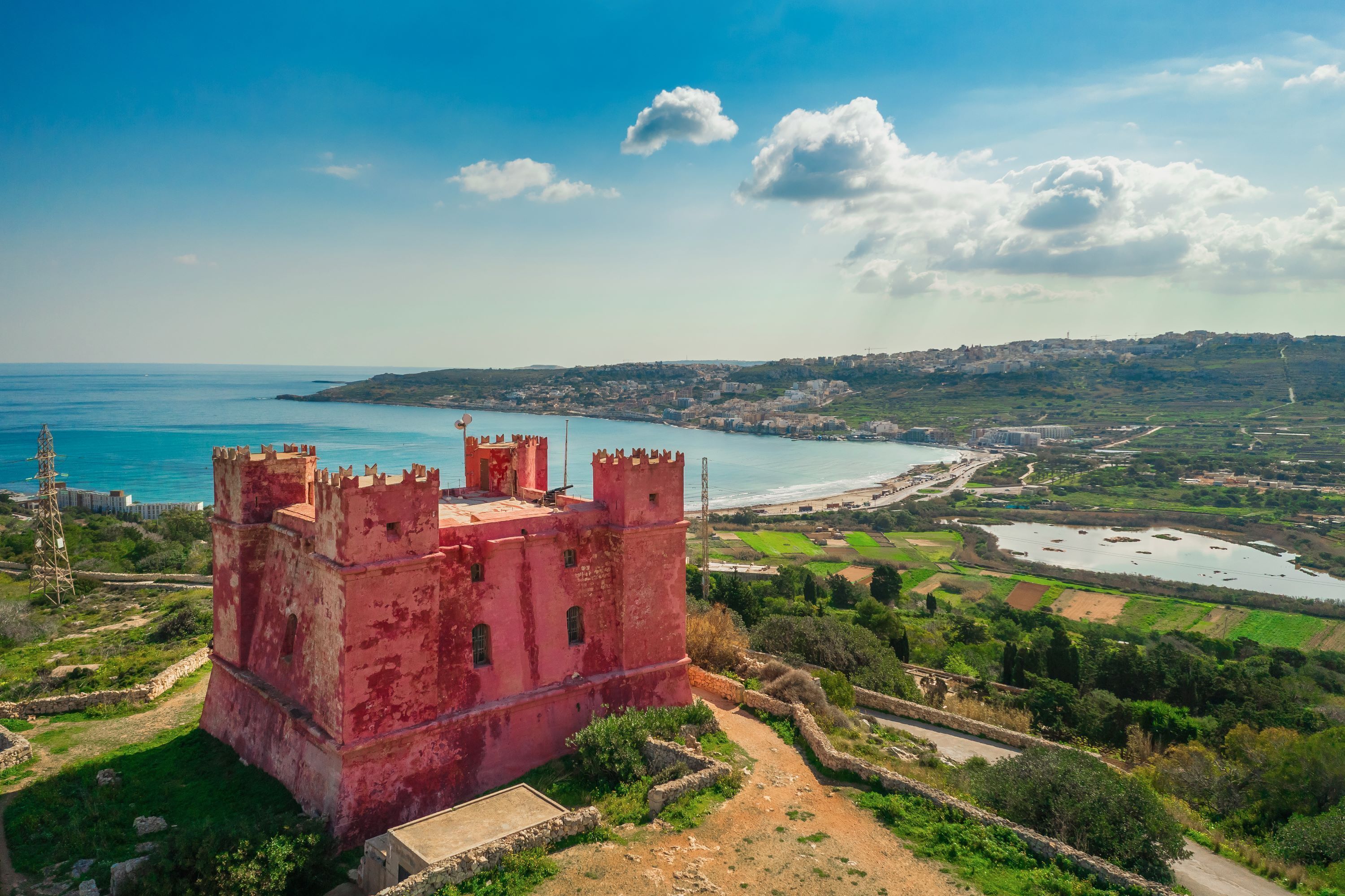 Une structure rouge, ressemblant à un château, d’où l’on a une vue dégagée sur le paysage environnant, notamment sur une étendue d’eau et des collines lointaines.
