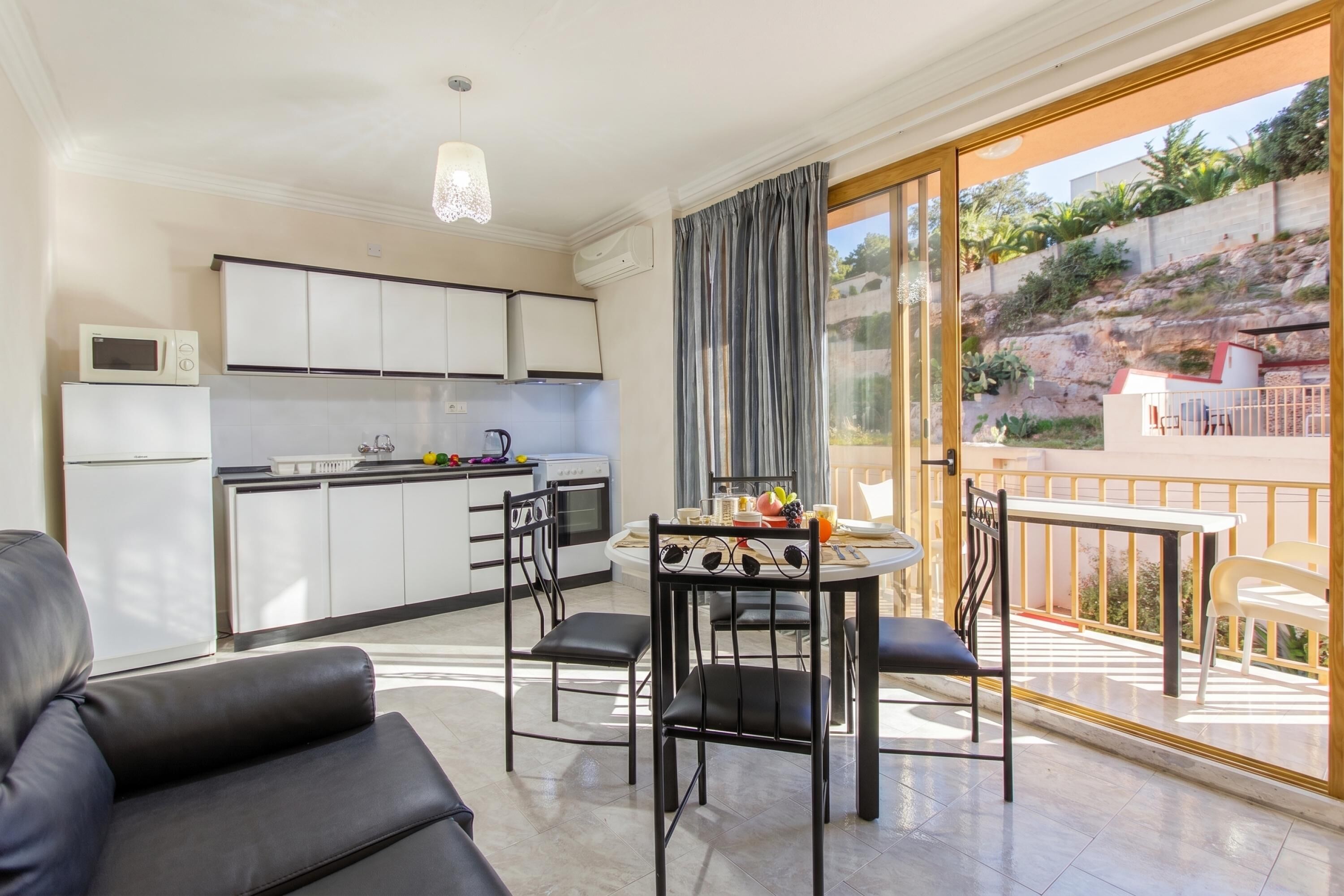 A modern kitchen with white cabinets, a microwave, and a kettle. There is a dining area with a table and chairs, and a balcony with a view of a rocky hillside.