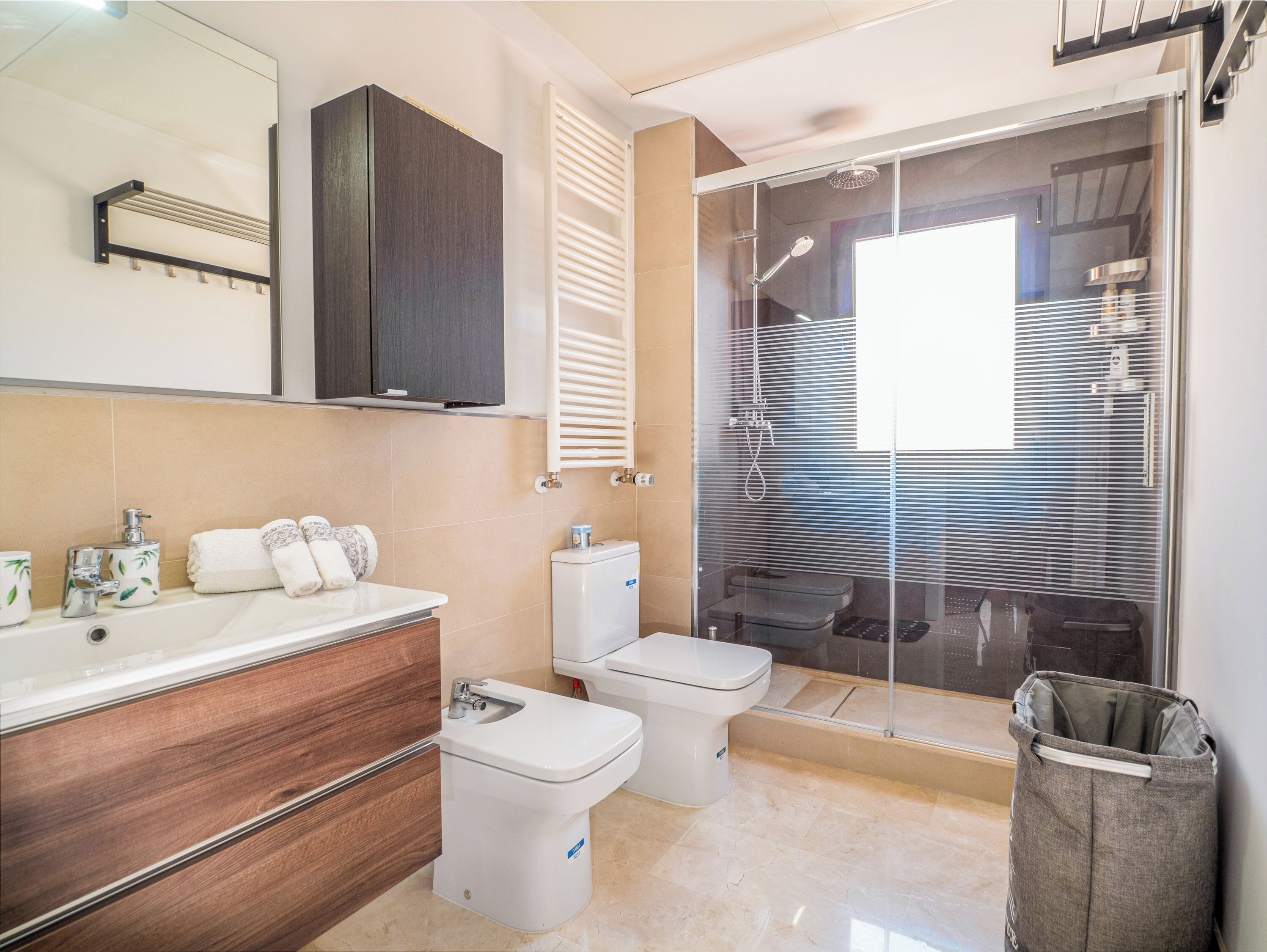 A modern bathroom with a wooden vanity, a white toilet, a glass-enclosed shower, and a towel rack.