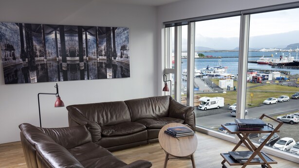 Een moderne woonkamer met een bruine leren bank, een klein houten bijzettafeltje en een groot raam met uitzicht op een haven met boten en voertuigen.