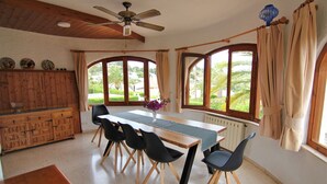 Zona pranzo con tavolo in legno, sedie nere, ventilatore a soffitto e ampie finestre con tende.