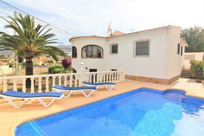 A swimming pool with lounge chairs, a white building with a tiled roof, and a palm tree.